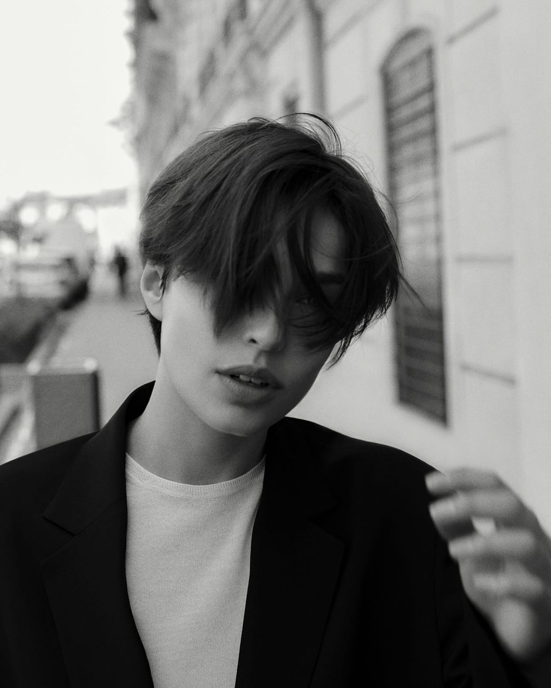Close-up black and white portrait of a person with short dark hair wearing a dark blazer over a white t-shirt on a city street.