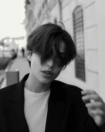 Close-up black and white portrait of a person with short dark hair wearing a dark blazer over a white t-shirt on a city street.