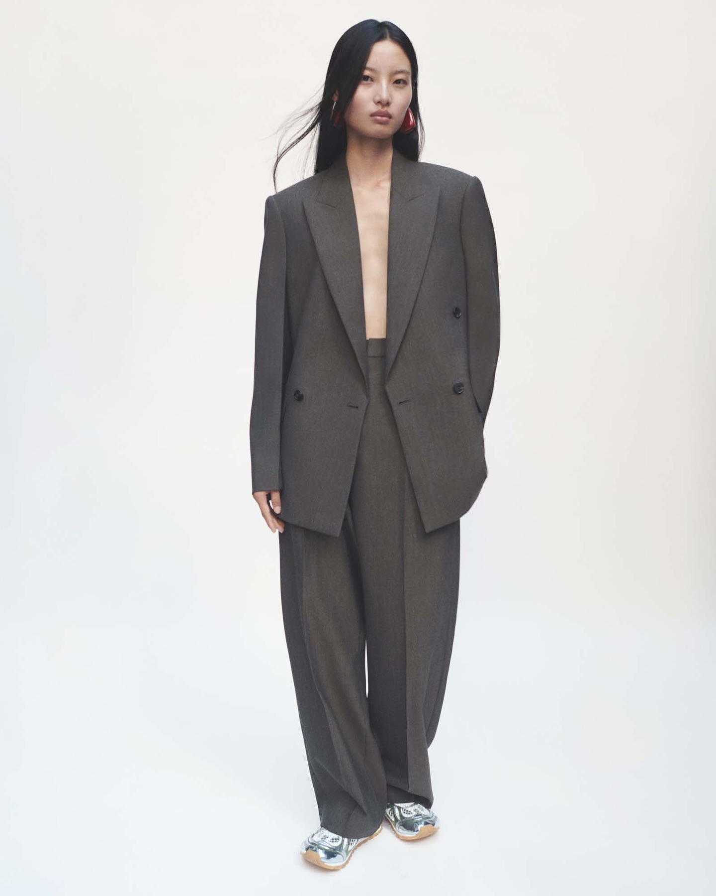 An Asian female fashion model wears a grey oversized suit and silver sneakers in a full shot against a white backdrop in a studio.