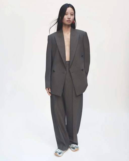 An Asian female fashion model wears a grey oversized suit and silver sneakers in a full shot against a white backdrop in a studio.