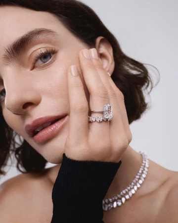 Close-up portrait of a woman wearing a large diamond ring and necklace, with her hand held to her face.