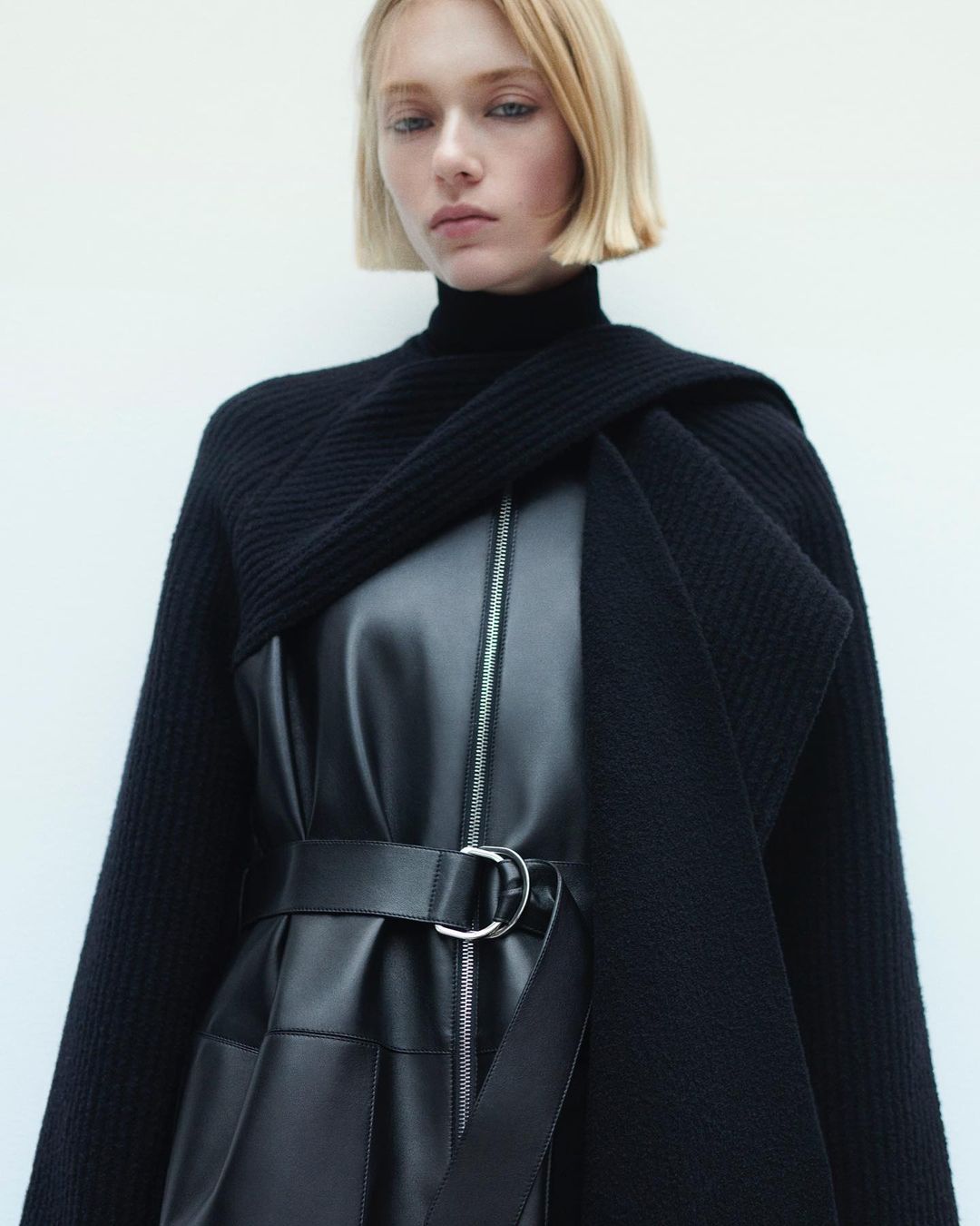 Fashion shot of a blonde woman wearing a black leather dress with a belt, sweater around the shoulders, set against a white backdrop.