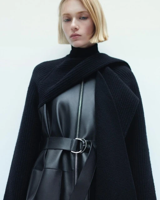 Fashion shot of a blonde woman wearing a black leather dress with a belt, sweater around the shoulders, set against a white backdrop.
