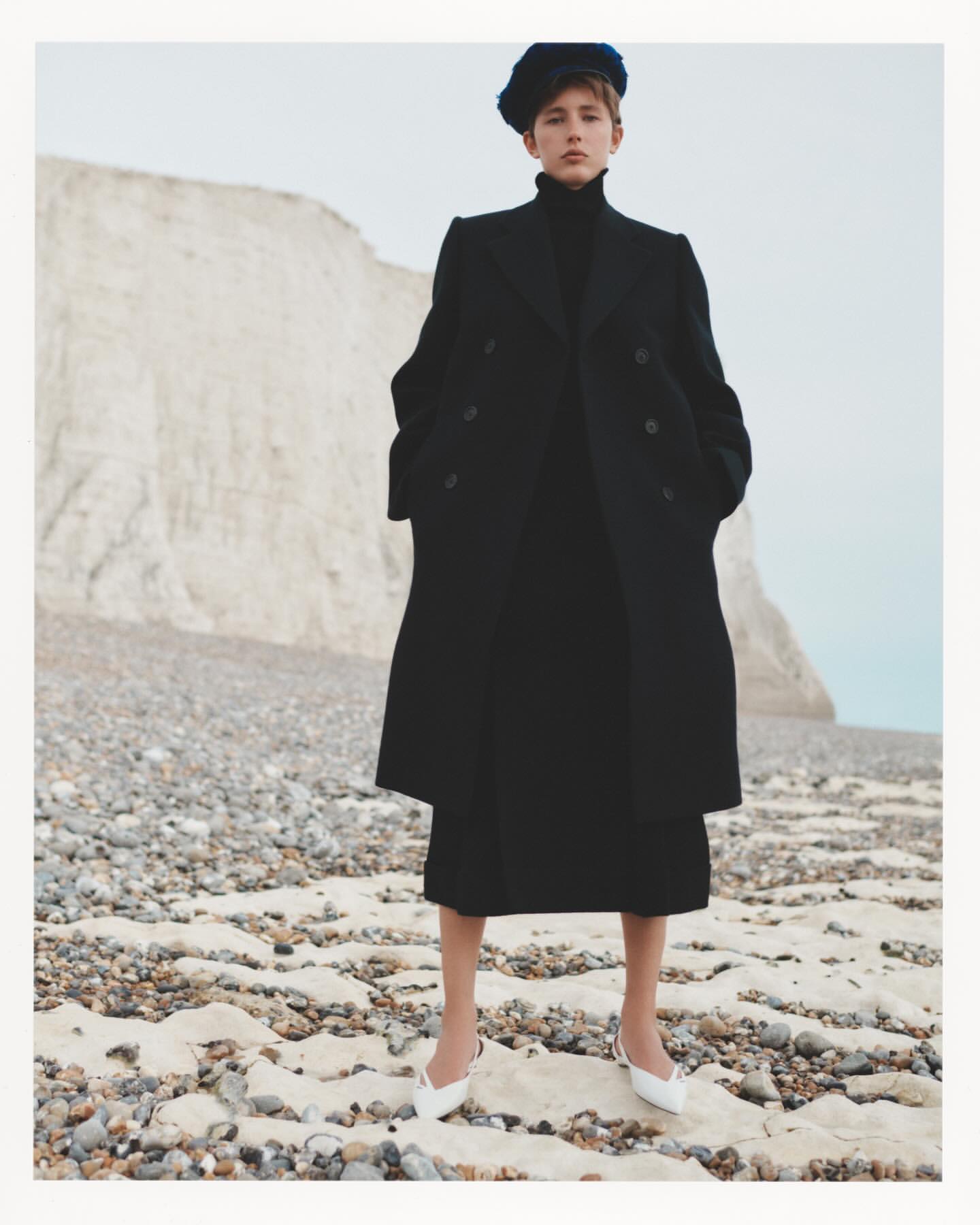 Woman in long black coat and blue hat poses on a rocky beach with white cliffs in the background, wearing white kitten heel shoes.