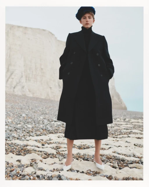 Woman in long black coat and blue hat poses on a rocky beach with white cliffs in the background, wearing white kitten heel shoes.