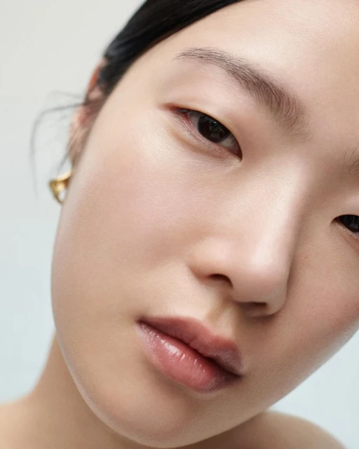 Close-up of Asian woman with neutral expression and glossy lips; minimal eye makeup and simple gold earrings in a minimalist style.