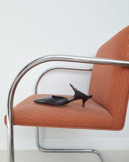Dark-brown heeled mule on an orange patterned chair with chrome frame in a brightly lit, neutral-toned interior space. 