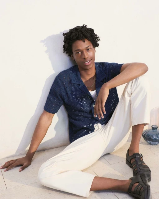 A young adult Black male poses indoors wearing a blue knit shirt and cream pants, with a serene expression and casual posture.
