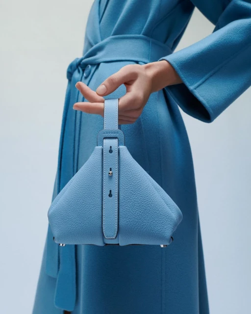 A powder-blue triangular handbag with an adjustable strap is held by a woman with a matching blue coat, creating a monochromatic look.
