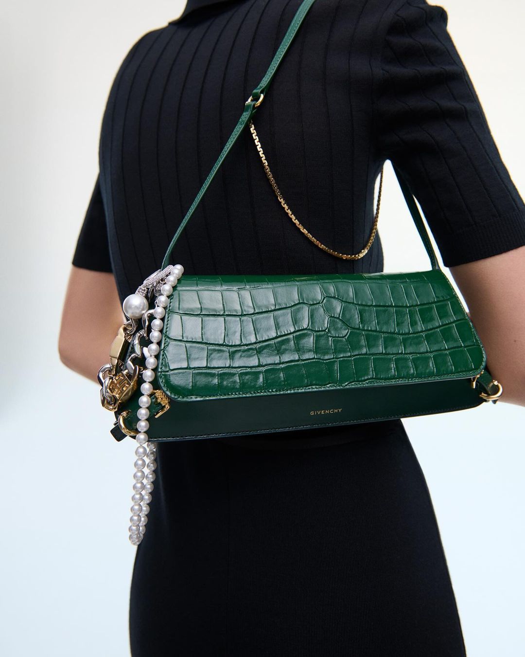 A woman wearing a black dress has a green crocodile-texture Givenchy bag over her shoulder with pearl and gold chain embellishments.