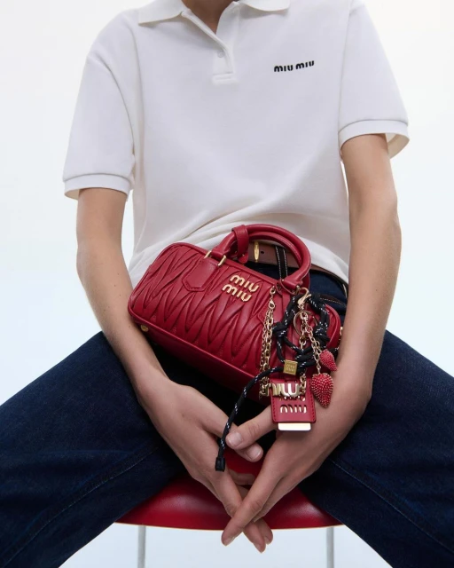 Person seated holding a red Miu Miu handbag. The bag has gold branding and a decorative keychain, against a clean, bright background.