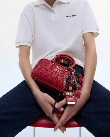 Person seated holding a red Miu Miu handbag. The bag has gold branding and a decorative keychain, against a clean, bright background.