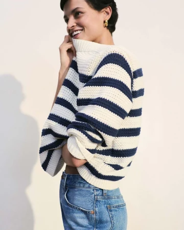 Woman wearing a striped sweater and blue jeans smiling against a plain white background