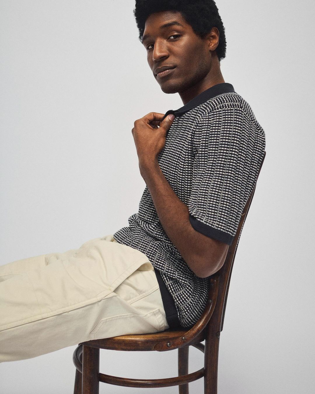 Dark-haired man in polo shirt sitting on chair against light background. Fashion-forward pose and studio lighting highlight the garment.