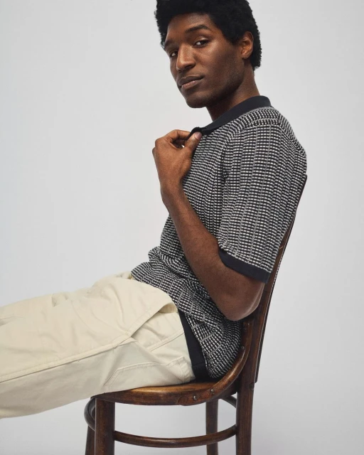 Dark-haired man in polo shirt sitting on chair against light background. Fashion-forward pose and studio lighting highlight the garment.