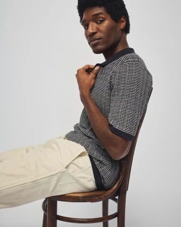 Dark-haired man in polo shirt sitting on chair against light background. Fashion-forward pose and studio lighting highlight the garment.