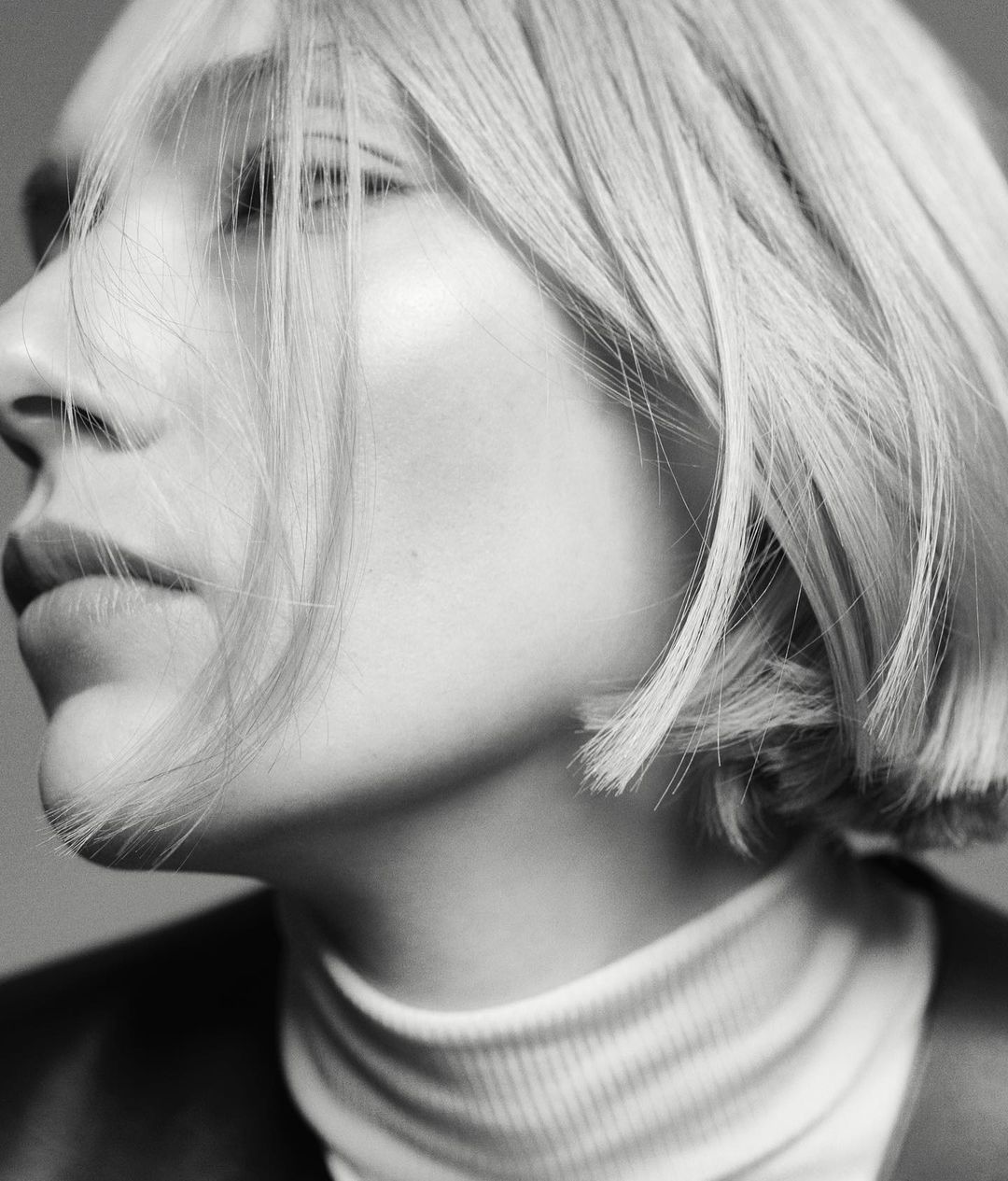 Black-and-white portrait of a fair-skinned woman with blonde bob, wearing a white turtleneck and black jacket, looking off-camera.