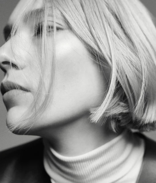 Black-and-white portrait of a fair-skinned woman with blonde bob, wearing a white turtleneck and black jacket, looking off-camera.