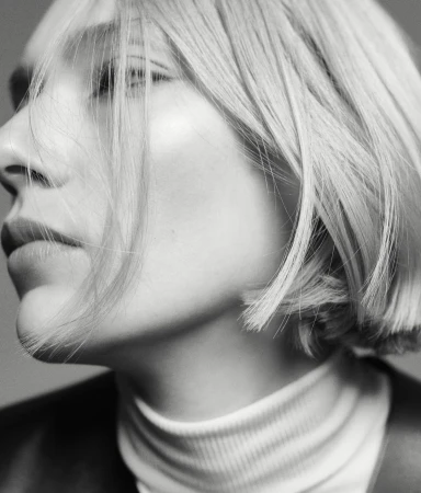 Black-and-white portrait of a fair-skinned woman with blonde bob, wearing a white turtleneck and black jacket, looking off-camera.