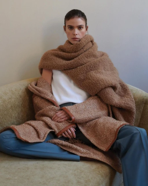 A woman sits on a couch, wrapped in a brown textured robe/scarf. She has blue pants and a white shirt. Neutral wall background.