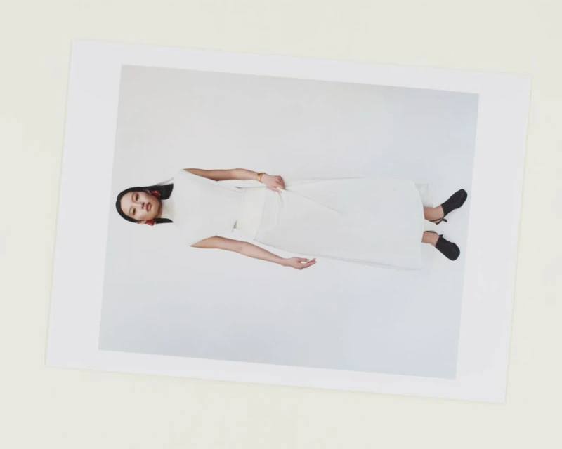 A woman in a white dress and black shoes is posing on a white background, showcased within a larger white frame, presented in a studio shot.