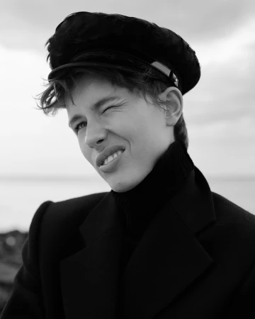 Monochrome portrait of a person wearing a dark cap and jacket, winking playfully against an overcast sky.