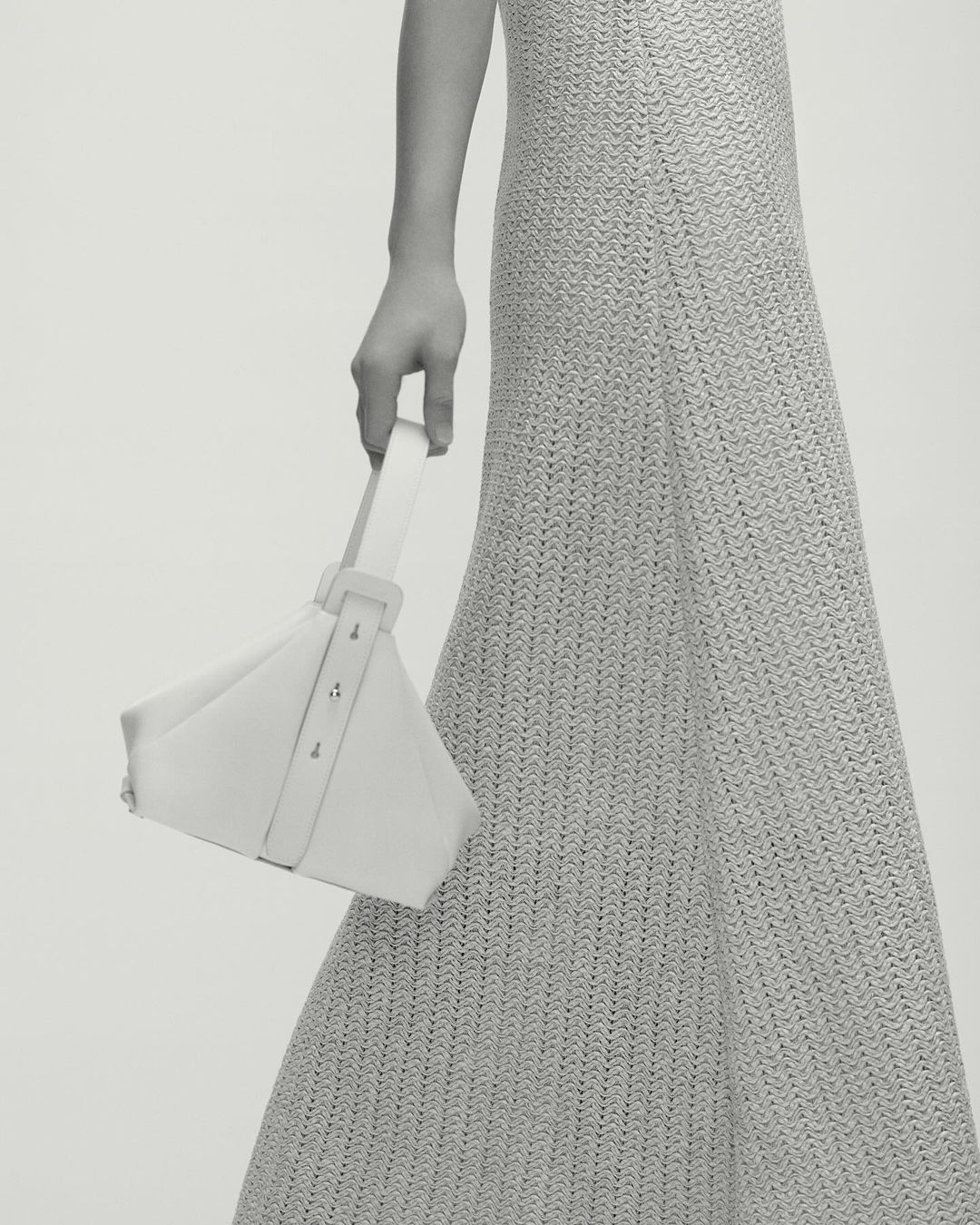 Fashion shot of a woman in a neutral dress holding a geometric white purse, showcasing minimalist style and texture against a plain background.