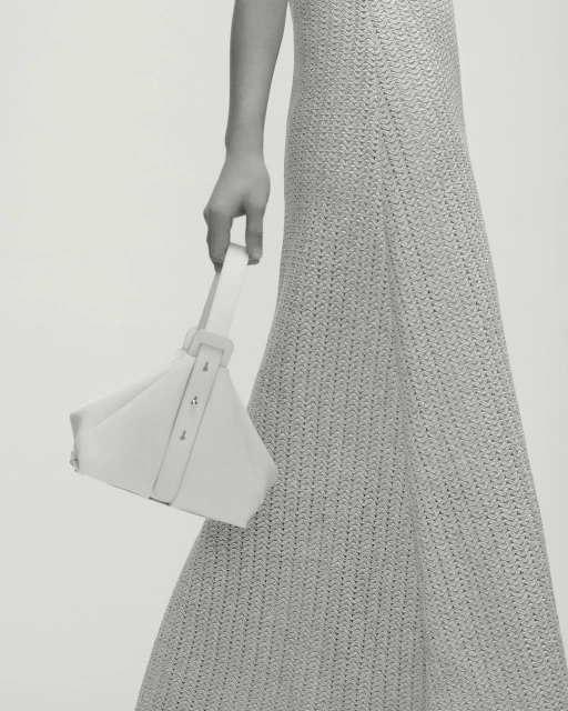 Fashion shot of a woman in a neutral dress holding a geometric white purse, showcasing minimalist style and texture against a plain background.