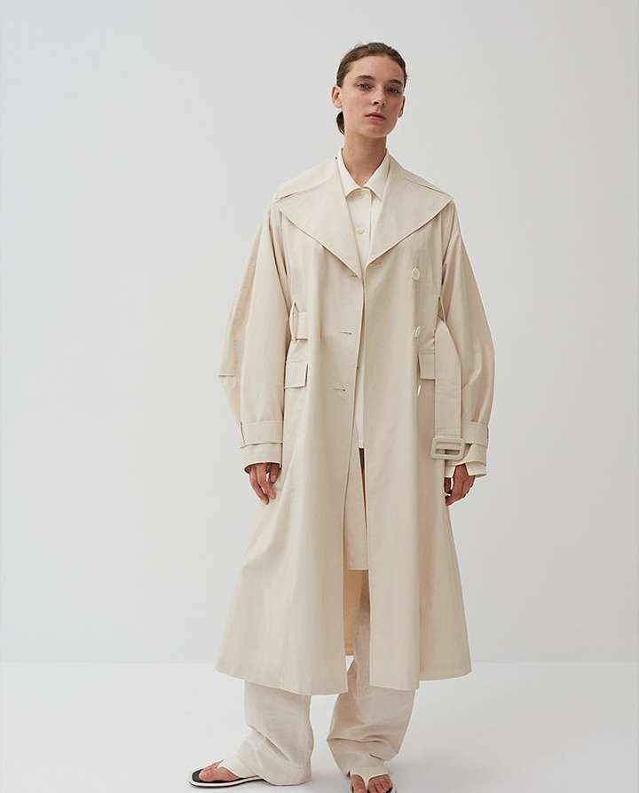A woman in a tan trench coat stands in a studio with neutral background. The coat has wide sleeves and a belt. She also wears pants and sandals.