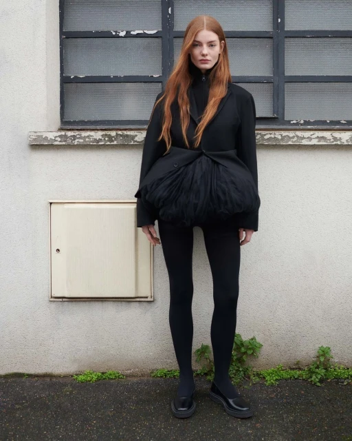 Red-haired woman in a black jacket with tulle detail, black leggings, and patent shoes, standing outdoors against a textured wall.