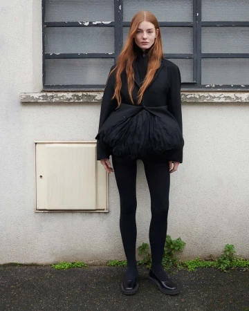 Red-haired woman in a black jacket with tulle detail, black leggings, and patent shoes, standing outdoors against a textured wall.
