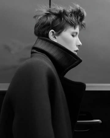 Black and white portrait of a person with short tousled hair wearing a dark high-collared coat