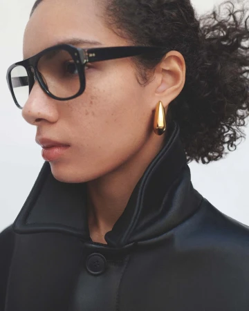 Close-up profile portrait of a model wearing thick black framed glasses and gold teardrop earrings in a black jacket