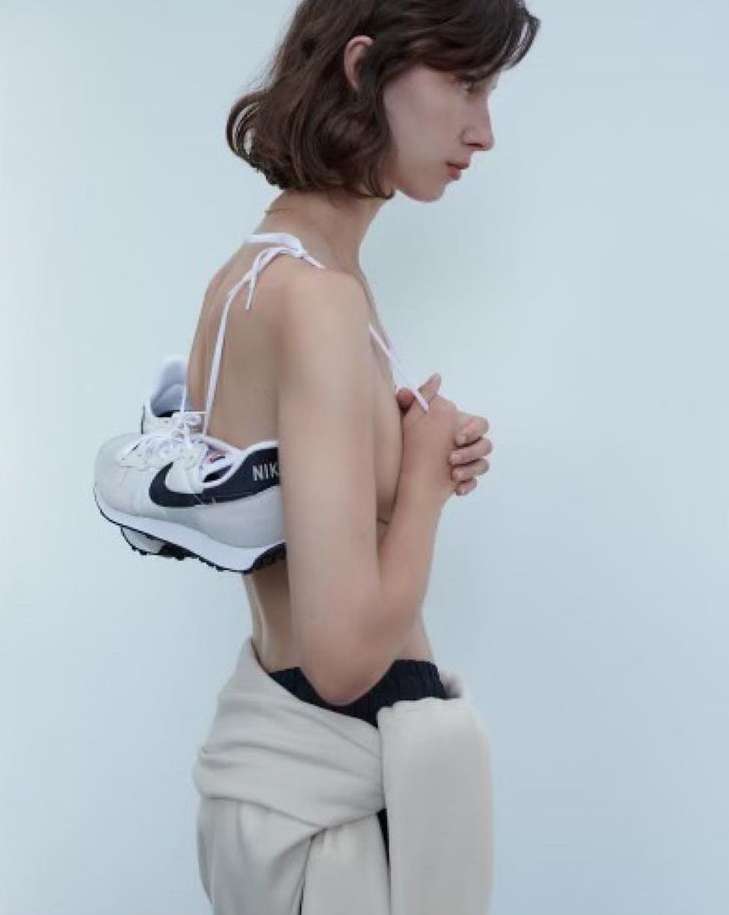 Young woman in profile with sneaker over her shoulder, wearing a draped neutral jacket, against a plain white background.