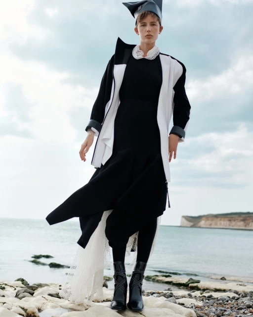 Fashion shot of a woman in a black dress with a white and black jacket and a gray hat posing on a rocky beach with cliffs in the background.
