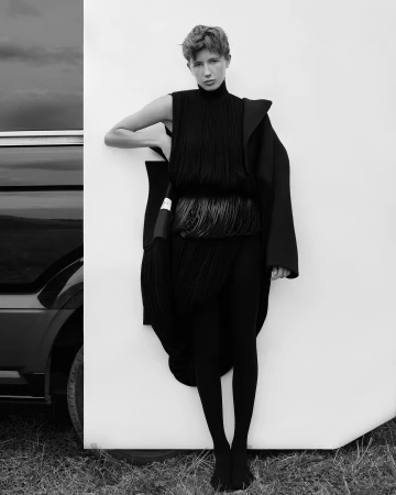 A slender woman poses with a black ensemble and short hair, leaning against a white backdrop next to a luxury car on a grassy surface.