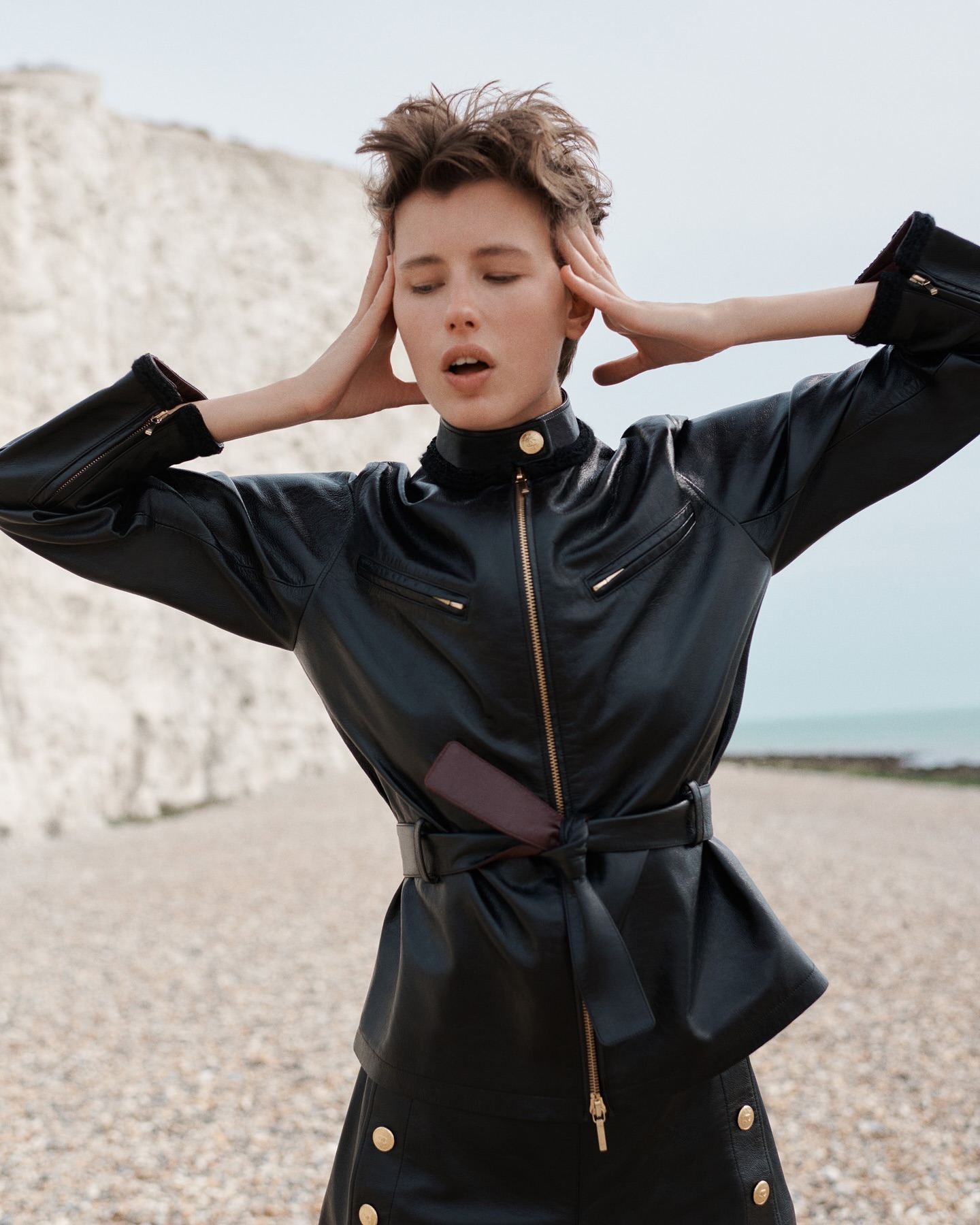 A young adult female with hands on her head poses on a beach wearing a black leather jacket and skirt with a light sky and chalk cliffs background.