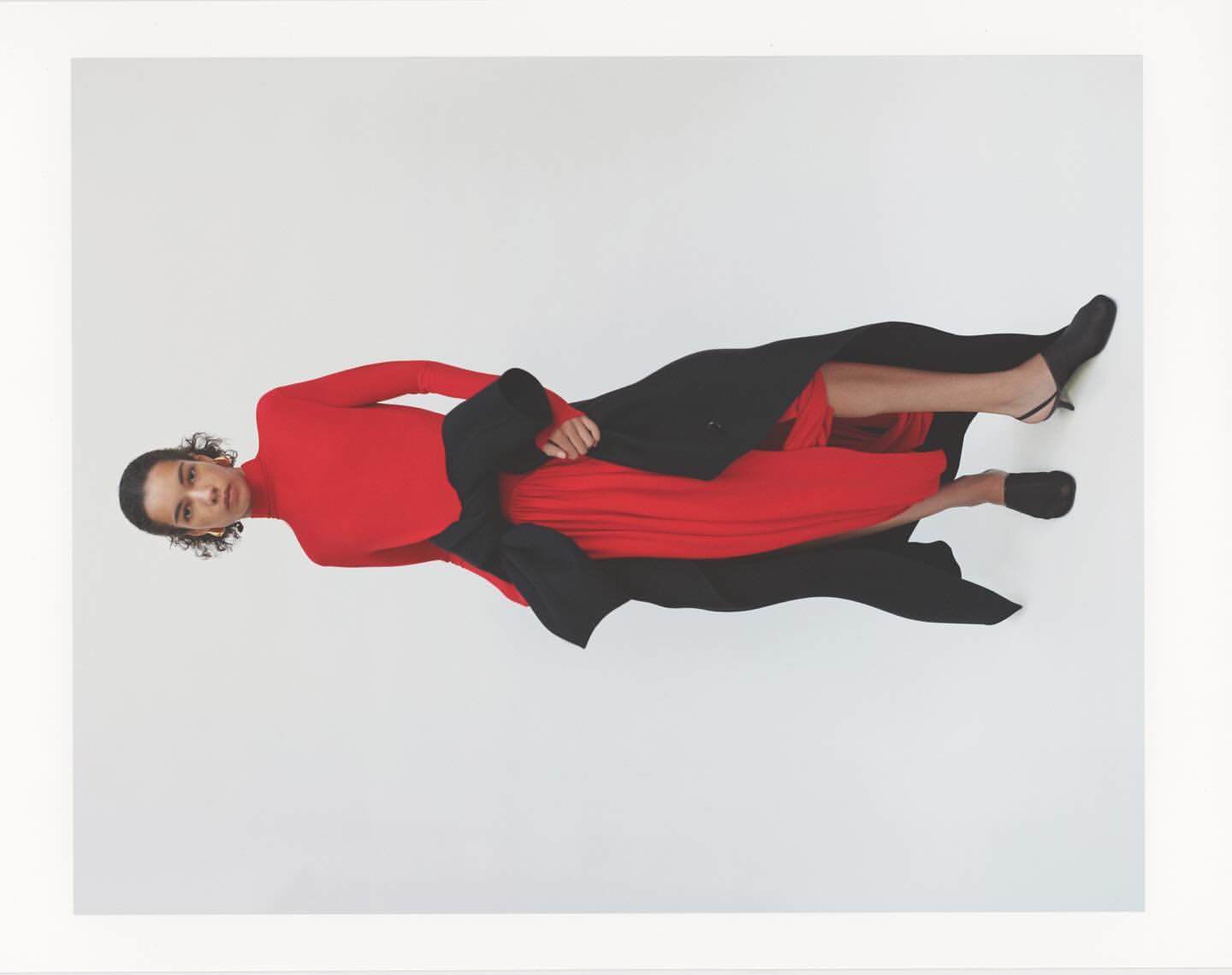 Model wearing a vibrant red turtleneck top and pleated red skirt paired with a dark coat against a plain white background.