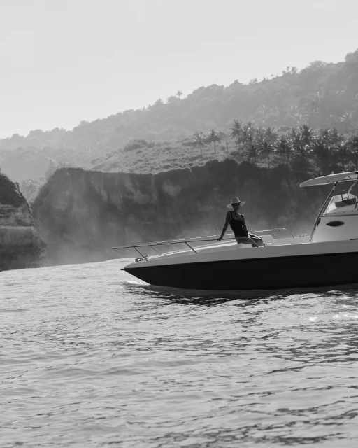 Woman in hat sits on bow of powerboat underway on ocean near cliffs and green hills in monochrome. Travel photography.
