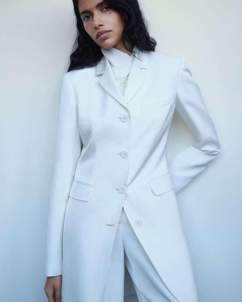 An adult woman is posing in a long white suit jacket and pants in a brightly lit indoor studio with a plain white backdrop.