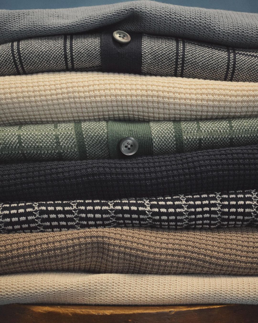 Pile of folded knitwear sweaters in neutral colors, showcasing various textures and patterns, with a plain dark background.
