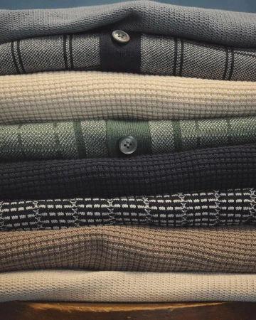 Pile of folded knitwear sweaters in neutral colors, showcasing various textures and patterns, with a plain dark background.