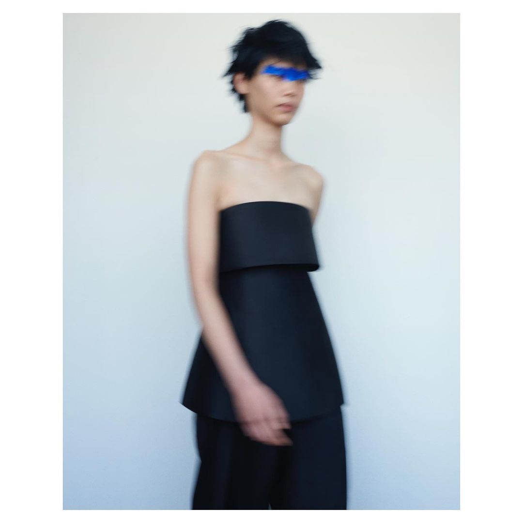 Avant-garde fashion photo featuring a slender figure in a black strapless top and matching pants against a stark white backdrop with blue paint over the eyes.
