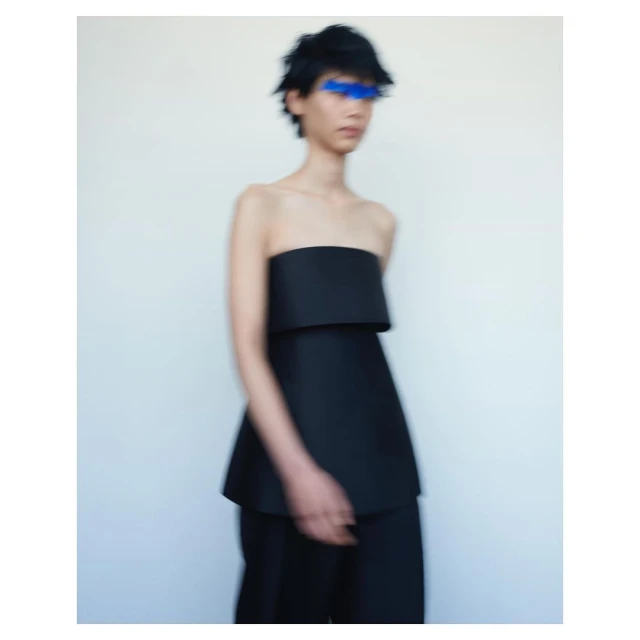 Avant-garde fashion photo featuring a slender figure in a black strapless top and matching pants against a stark white backdrop with blue paint over the eyes.