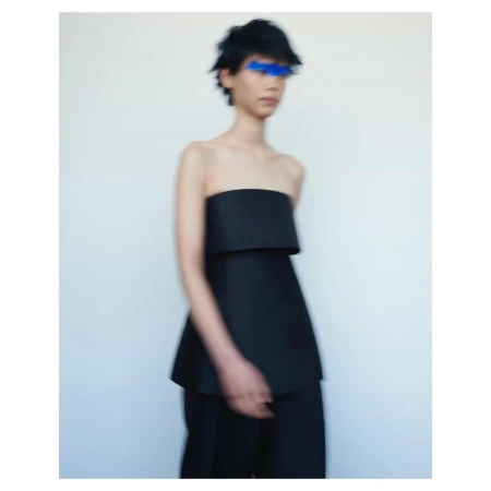Avant-garde fashion photo featuring a slender figure in a black strapless top and matching pants against a stark white backdrop with blue paint over the eyes.