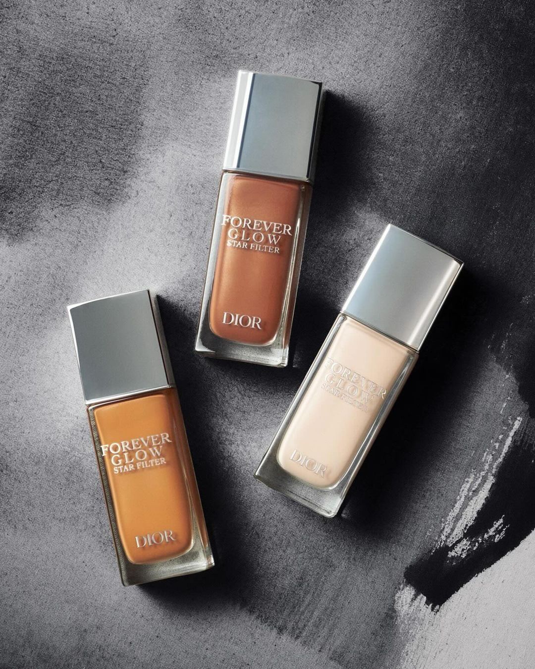 Three bottles of Dior Forever Glow Star Filter foundation are displayed against a textured gray background, showcasing different shades.