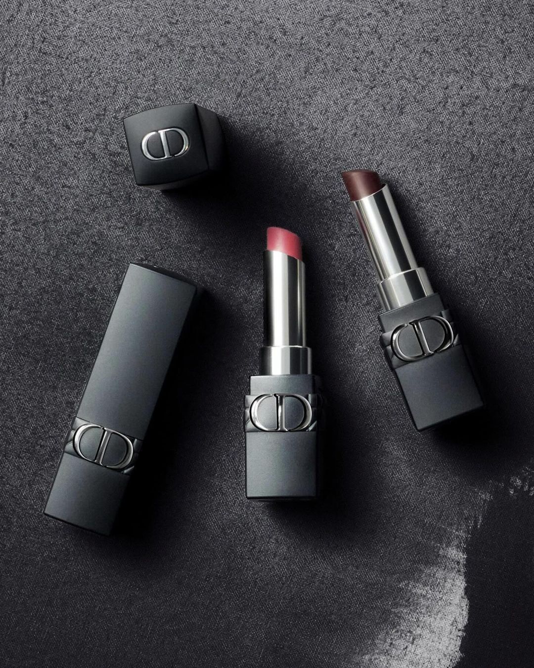 Elevated studio shot of three Dior lipsticks with matte black cases and silver accents on a dark gray textured background. 