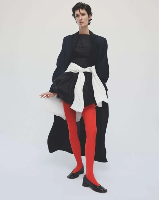 Fashion shot of a person with short dark hair in black top, dress with white bow, red tights, black flats and navy coat on white backdrop.