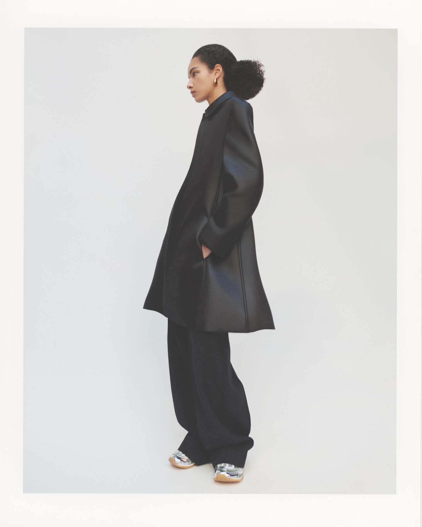 Model wearing a dark oversized coat and trousers against a clean white seamless backdrop