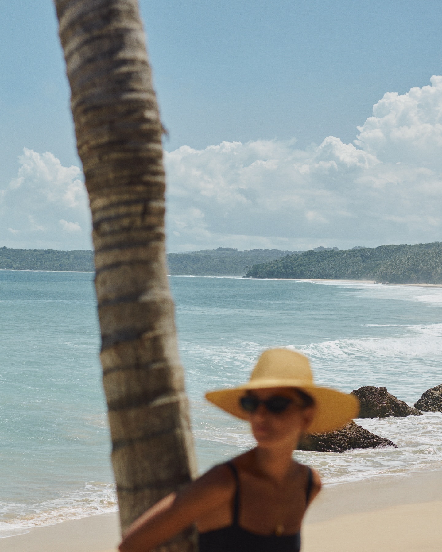 Woman in a straw hat stands near a palm tree on a sunny beach with a turquoise sea and distant islands in the background.